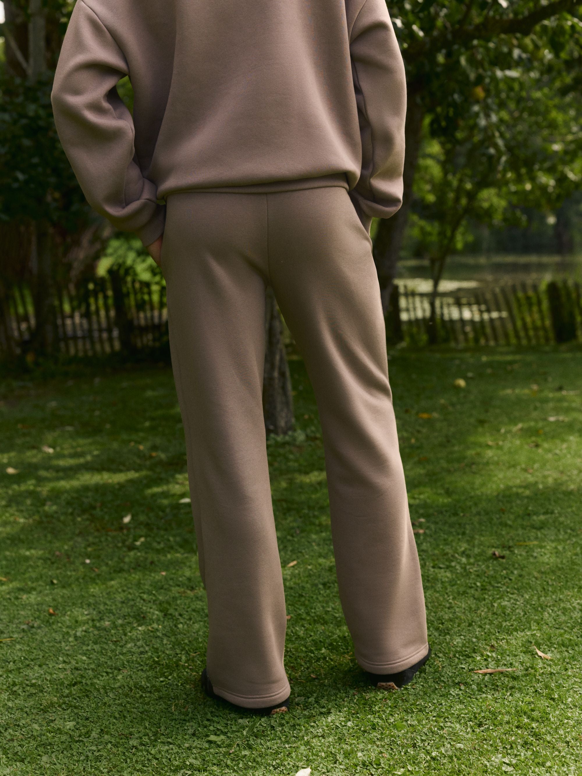 Man wearing walnut CASPARA straight-leg sweatpants standing on a grassy lawn, seen from behind.